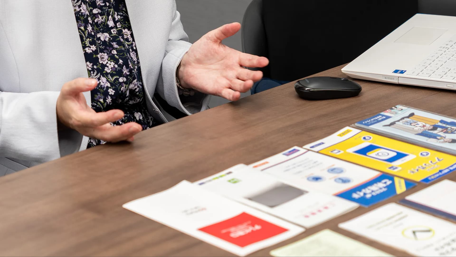 写真。胸から下に映ってる監修者と思わしき人が、テーブルに並べたれたパンフレットを元に手振りを交えながら話をしている様子